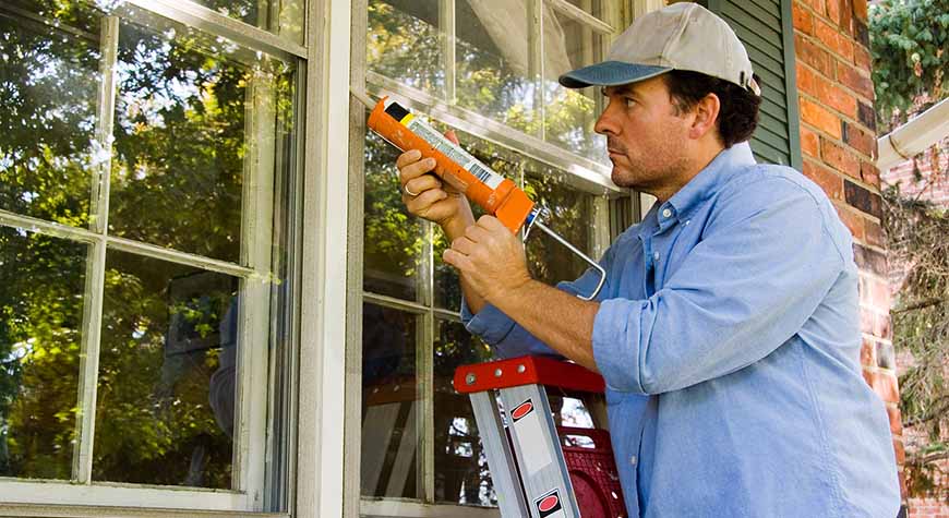 man on stepladder caulks his windows